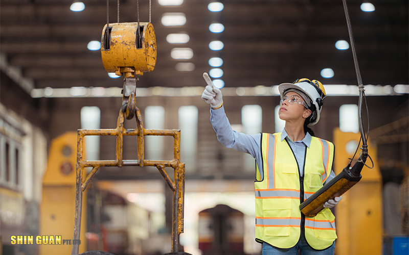 Worker in safety gear operating an overhead crane and inspecting the lifting hook inside an industrial facilit